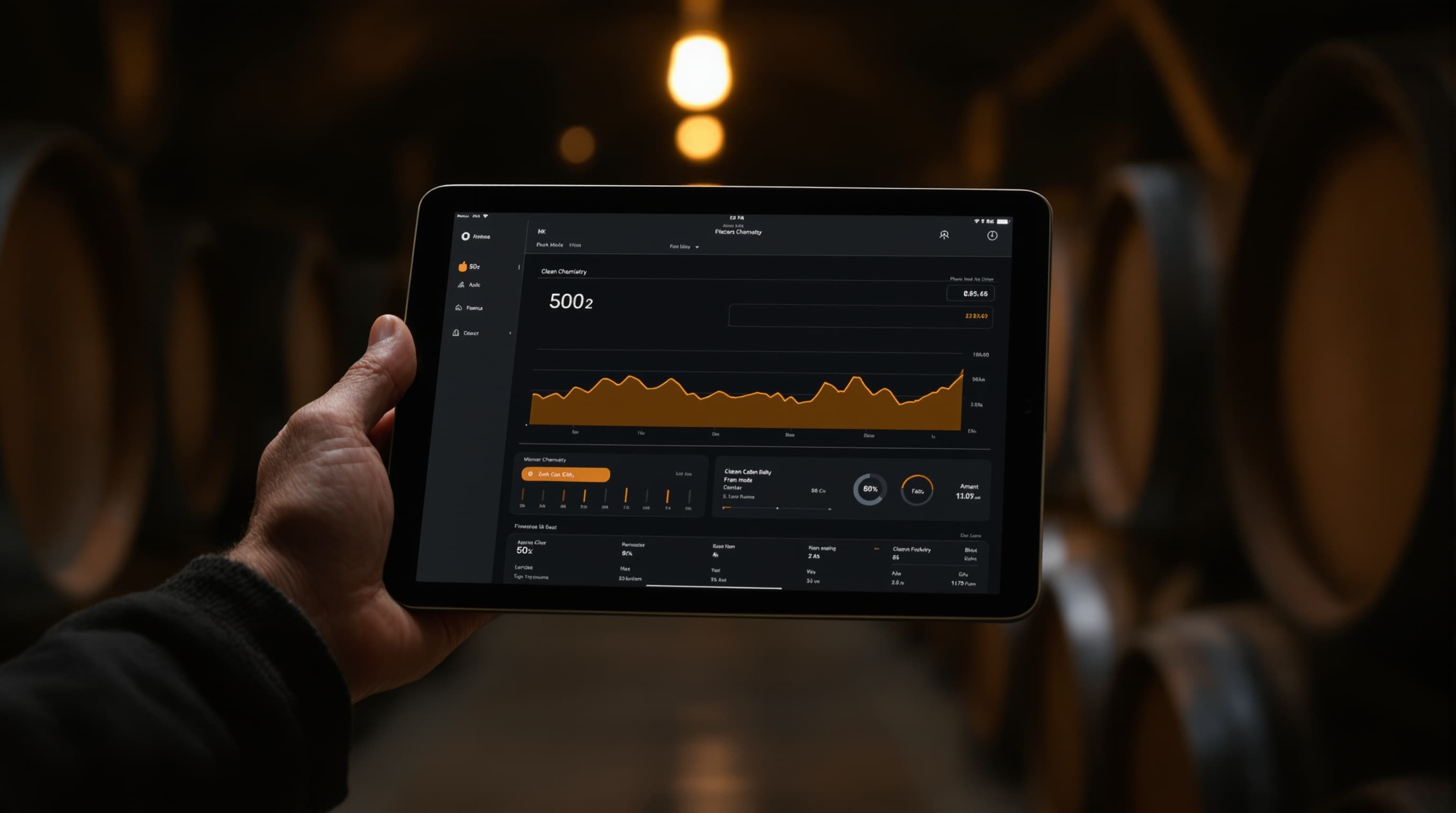 A winemaker's hand holding a tablet showing a dark-mode chemistry dashboard with an SO₂ trend line, dim winery cellar behind